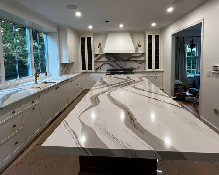 A long kitchen island in white Cambria quartz with bold gray and gold veins, white cabinets and tall windows behind it