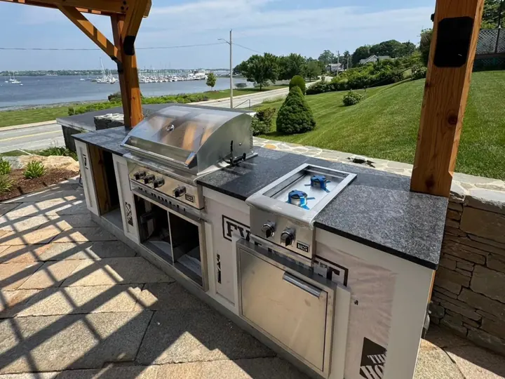 Outdoor kitchen under a wooden pergola, grills set into leathered Steel Gray granite, the water behind